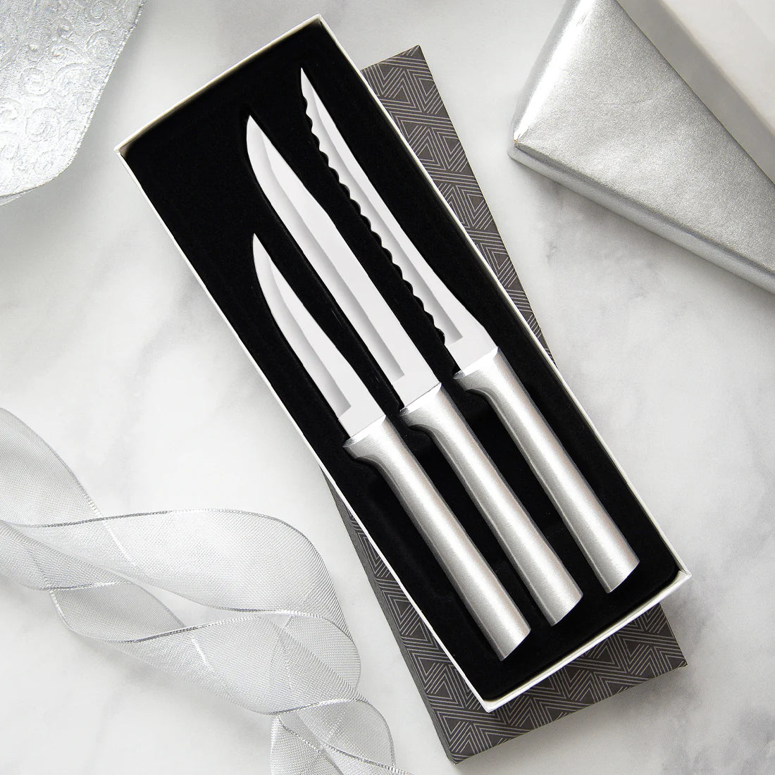 Three stainless steel kitchen knives with silver handles, including a steak knife and two utility knives, presented in an open black gift box on a light marble surface with decorative ribbon and a silver box lid.