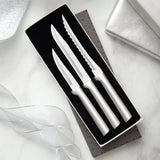 Three stainless steel kitchen knives with silver handles, including a steak knife and two utility knives, presented in an open black gift box on a light marble surface with decorative ribbon and a silver box lid.