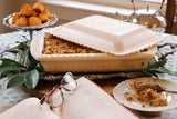 A white ceramic foil casserole dish on a table, partially covered with a lid, with food inside. There is a plate with a piece of the food, cutlery, and a notebook with a pen on the table.