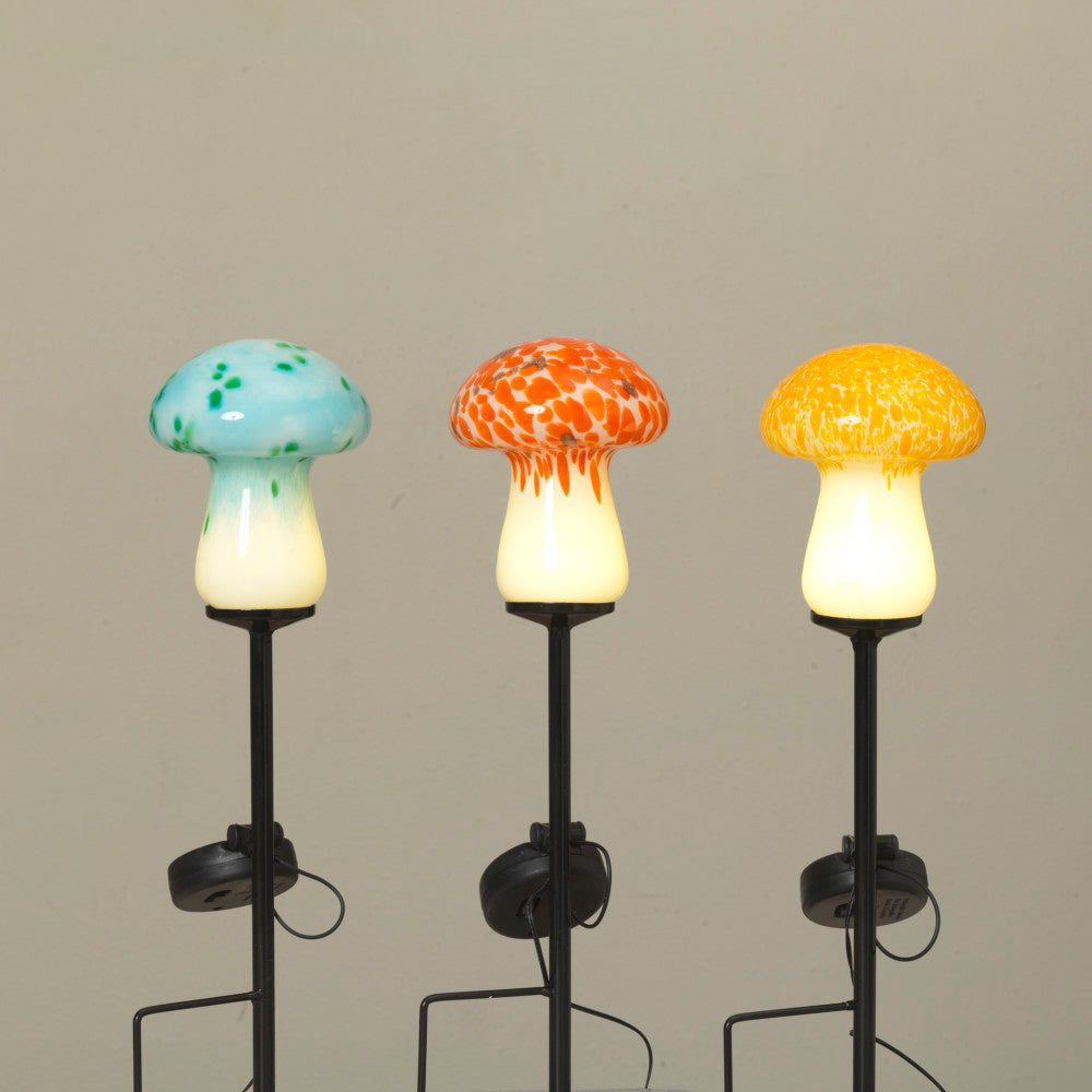This image displays a set of three solar lighted glass mushrooms, each distinctively colored to enhance visual appeal. The mushrooms are designed with a translucent glass top, available in shades of blue, orange, and yellow, complemented by a solid black stem.