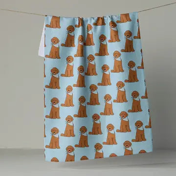 A kitchen towel with a repeating pattern of golden retriever dogs wearing white bandanas on a light blue background, hanging from a string against a neutral gray wall.