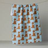 A kitchen towel with a repeating pattern of golden retriever dogs wearing white bandanas on a light blue background, hanging from a string against a neutral gray wall.