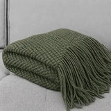 An olive green textured knit throw blanket with long fringe, folded and displayed on a light gray upholstered sofa.