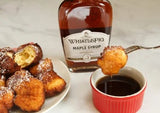 A bottle of WhistlePig Maple Syrup with a plate of pancakes drizzled with syrup and a small red bowl of syrup on a white surface. A spoon with syrup on it is also visible.