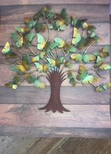 A wall-mounted metal tree decor with green and yellow leaves on a brown tree trunk, placed against a wooden background.