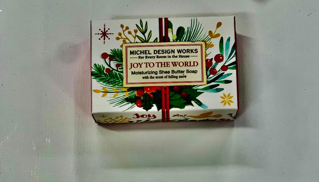 This image features a packaged bar of soap, specifically from Michel Design Works. The bar is contained within a brightly colored box that is decorated with a festive, Christmas-themed design, including holly and other holiday greens.