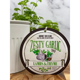 A packaged gourmet herb dip blend labeled 'Zesty Garlic Dip' by Lambs & Thyme, displayed on a wooden surface with green herbs in the background.