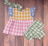 A set of baby clothing including a pinafore dress with yellow, blue, and red checkered patterns, accompanied by a green and white checkered bow headband and matching bloomers.