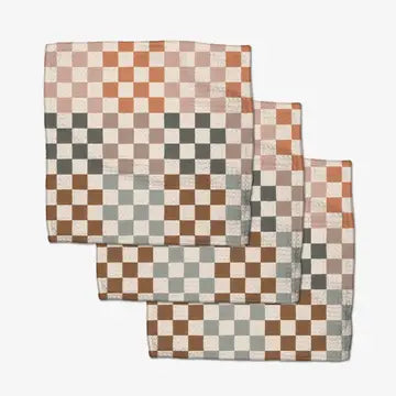 Three square checkered cloth napkins in earthy tones of terracotta, brown, sage green, and cream, arranged in a cascading stack on a light gray background.