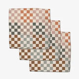 Three square checkered cloth napkins in earthy tones of terracotta, brown, sage green, and cream, arranged in a cascading stack on a light gray background.