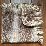 A beige and brown faux fur blanket with a satin ruffled trim and a matching ruffled pillow cover, laid flat on a wooden floor.