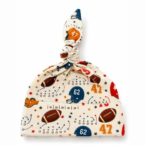 A baby knit hat with a top knot, featuring a sports-themed print of footballs, helmets, stars, and numbers in red, blue, and brown on a cream background.