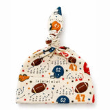 A baby knit hat with a top knot, featuring a sports-themed print of footballs, helmets, stars, and numbers in red, blue, and brown on a cream background.