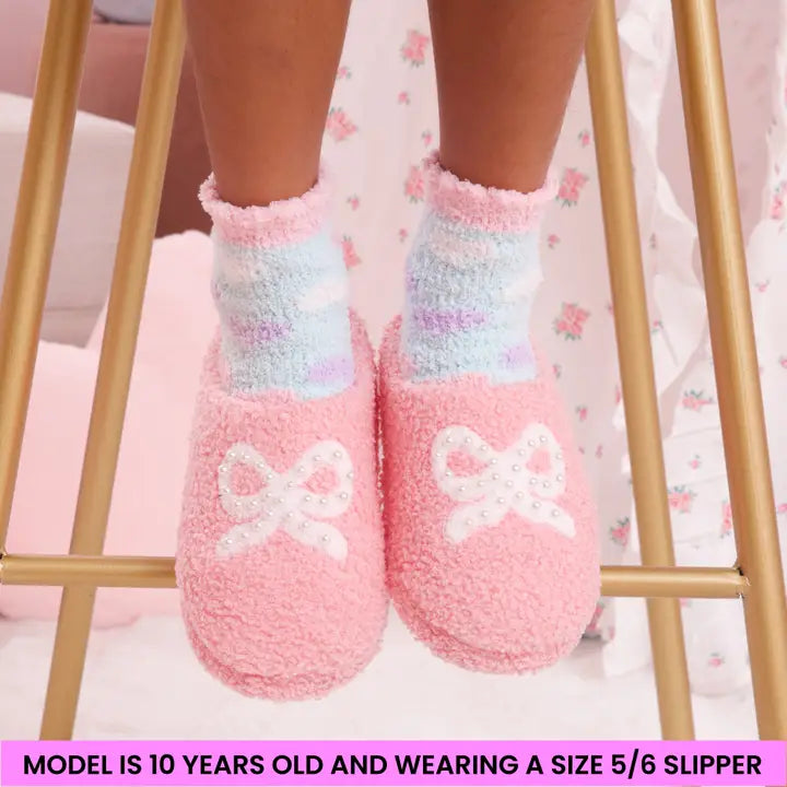 Pink fuzzy slippers with a bow worn by a child, sitting on a gold stool.