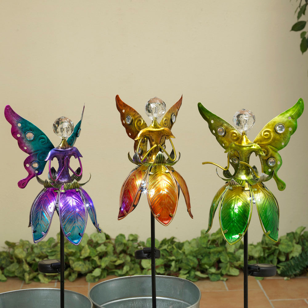 Three solar lighted fused glass fairies in green, purple, and yellow colors, standing on stakes with a plant in the background.