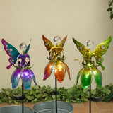 Three solar lighted fused glass fairies in green, purple, and yellow colors, standing on stakes with a plant in the background.