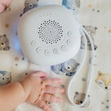 A white baby sleep monitor or humidifier with buttons and a wrist strap, placed on a baby blanket with teddy bear prints. A baby's hand is resting near the device.