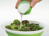 A collapsible green salad bowl with a person's hand pouring salad dressing into it from a squeeze bottle.