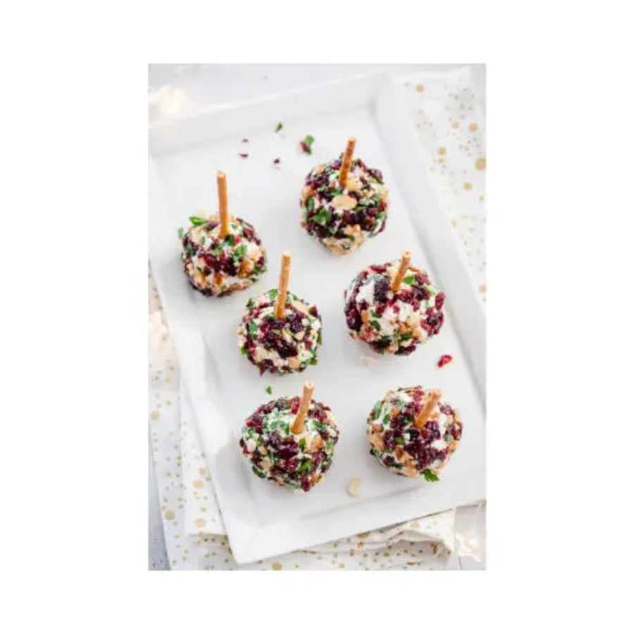Dessert balls with cranberries and nuts on skewers on a white surface.