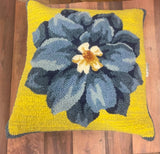 A square-shaped throw pillow with a blue hooked flower design on a yellow background, placed on a wooden floor.
