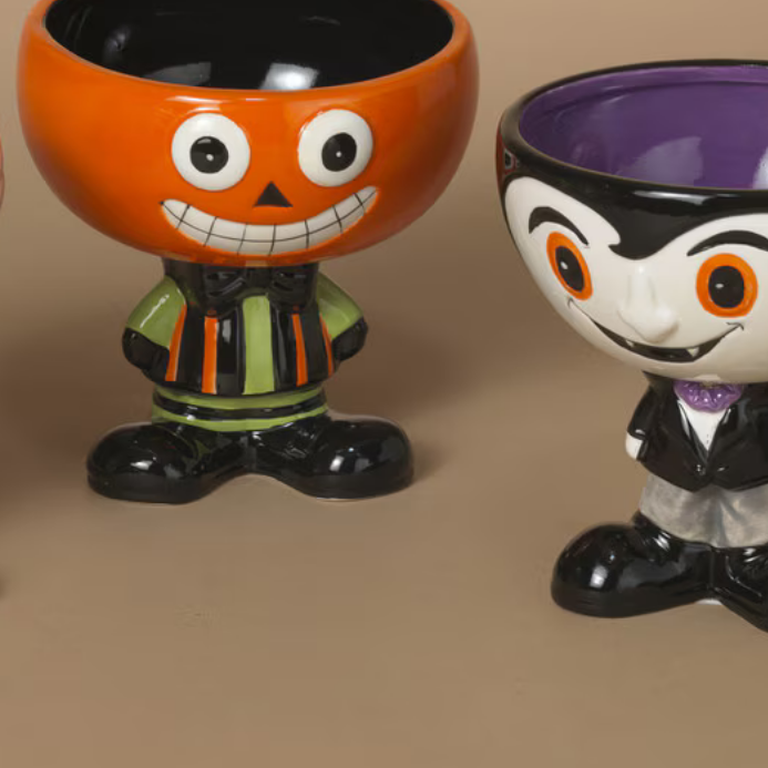 Two decorative bowls with Halloween designs, one in the shape of a smiling pumpkin and the other with a character resembling a trick or treat bag, both with a black base and white details.