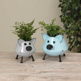 Two metal planters designed to look like stylized pigs, one silver and one blue, with plants growing out of them, placed on a wooden surface with a plant in the background.