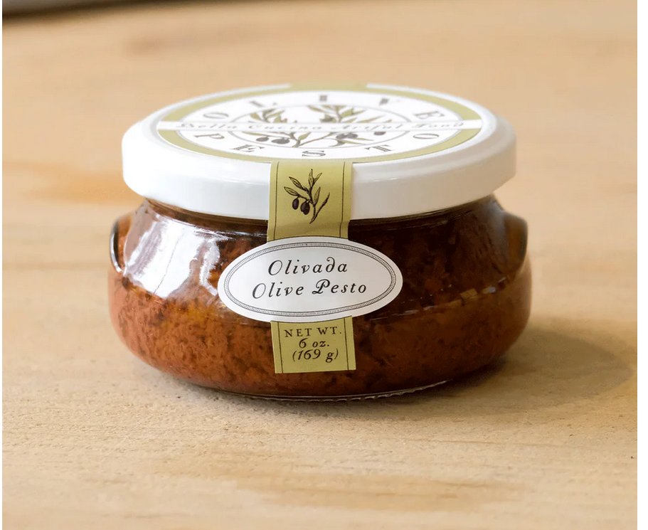 A jar of OLIVADA OLIVE PESTO with a label showing the brand name and the product name, placed on a wooden surface.