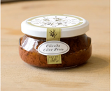 A jar of OLIVADA OLIVE PESTO with a label showing the brand name and the product name, placed on a wooden surface.