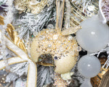 A close-up of a decorative holiday ornament shaped like a donut, covered in gold glitter, white pearls, and clear rhinestones, hanging on a white-frosted Christmas tree alongside gold ribbons and white glass ornaments.