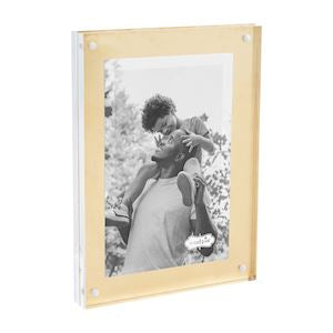 A brass acrylic picture frame with a photograph of a person embracing an animal. The frame has a clear glass front and a gold-colored metal border.