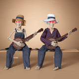 Two skeleton figures dressed in overalls, hats, and sunglasses, sitting and playing banjos.