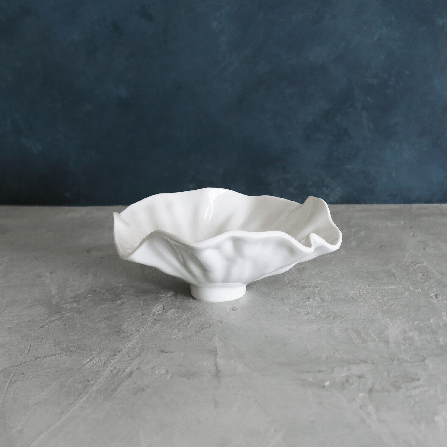 A white, ceramic VIDA Bloom small bowl with a petal-like rim, placed on a grey surface with a blue background.