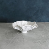 A white, ceramic VIDA Bloom small bowl with a petal-like rim, placed on a grey surface with a blue background.