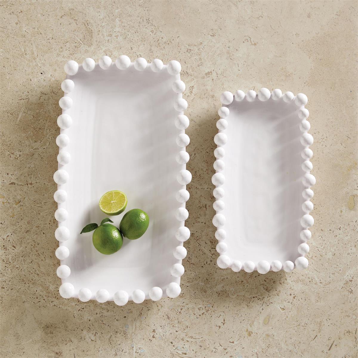 Two white ceramic serving platters with a dotted rim design, one large and one small, displayed on a sandy surface with some green olives on top of the larger platter.
