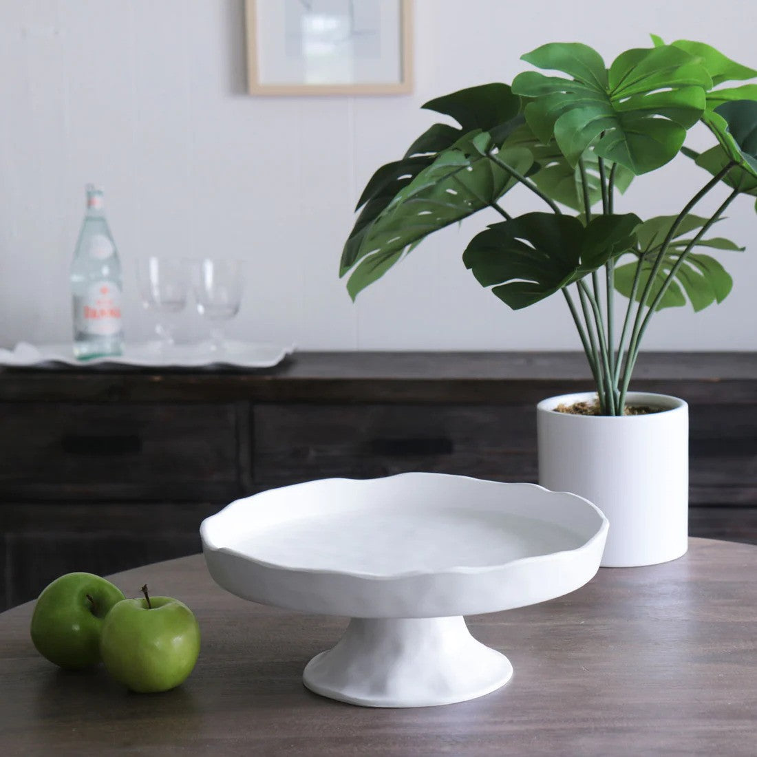 A white, round pedestal cake plate on a wooden table with a green plant in a white vase in the background.