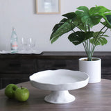 A white, round pedestal cake plate on a wooden table with a green plant in a white vase in the background.