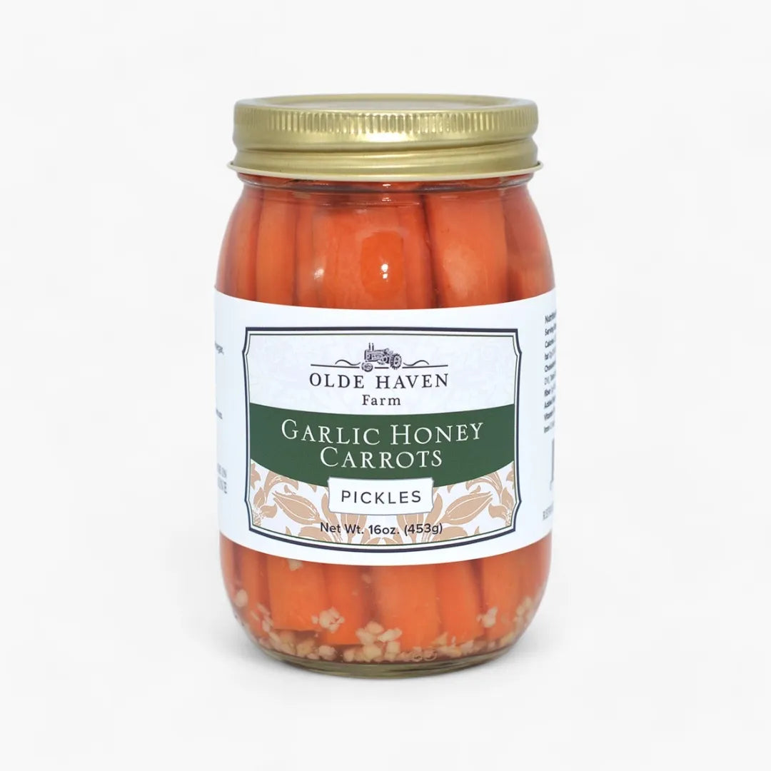 A jar of Olde Haven Farm Garlic Honey Carrots Pickles, featuring bright orange carrots in a glass jar with a gold metal lid and a white label with green and beige accents.