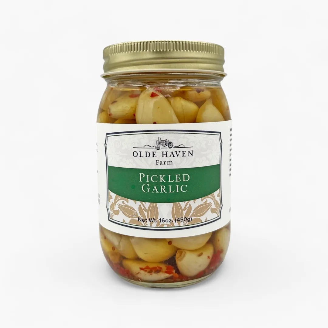 A jar of Olde Haven Farm Pickled Garlic on a clean white background. The glass jar contains pale yellow garlic cloves in a brine with visible red pepper flakes, sealed with a gold metal lid and featuring a white and green label.