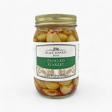 A jar of Olde Haven Farm Pickled Garlic on a clean white background. The glass jar contains pale yellow garlic cloves in a brine with visible red pepper flakes, sealed with a gold metal lid and featuring a white and green label.