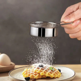 A stainless steel powered sugar sieve being used to sprinkle sugar over a waffle on a plate.