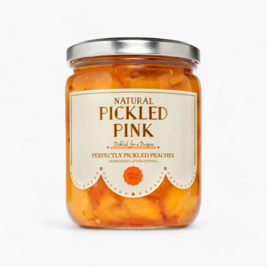 A glass jar of Pickled Pink Perfectly Pickled Peaches by Pickled by a Purpose, featuring a cream-colored label with brown text and a silver metal lid, set against a plain white background.