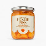 A glass jar of Pickled Pink Perfectly Pickled Peaches by Pickled by a Purpose, featuring a cream-colored label with brown text and a silver metal lid, set against a plain white background.