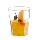 A clear glass cocktail ornament with a decorative metal tag, an orange slice, and a cherry on top, representing a miniature old fashioned cocktail.