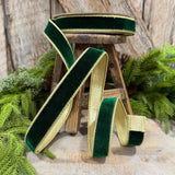 Dark green velvet ribbon with a gold glitter border, styled draped over a small wooden stool with green pine branches in the background.