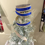 A decorative flocked Christmas tree with silver-white needles, wrapped with three blue and gold ribbons, in an indoor setting.