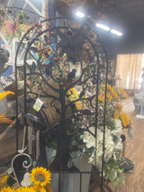 A metal arched garden trellis with a tree of life design, displayed in a garden center setting with various plants and flowers around it.