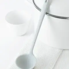 A white slim ladle is standing upright beside a white pot with a lid on a white surface, with a textured white mat below.