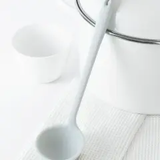 A white slim ladle is standing upright beside a white pot with a lid on a white surface, with a textured white mat below.