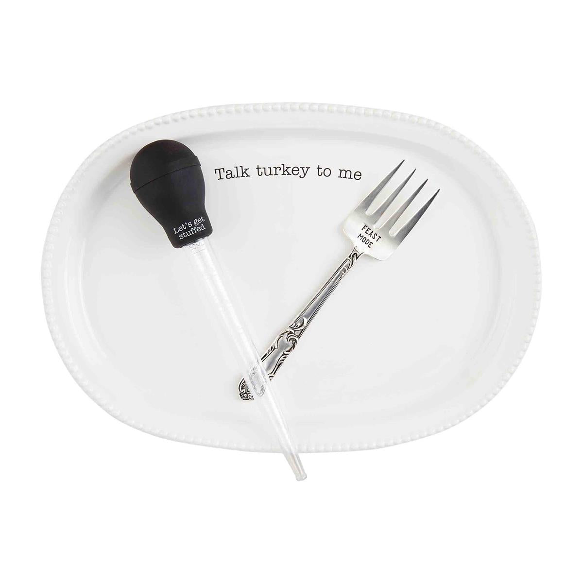 A white ceramic platter with the phrase 'Talk turkey to me' printed on it, accompanied by a silver gravy boat and a silver fork.