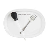 A white ceramic platter with the phrase 'Talk turkey to me' printed on it, accompanied by a silver gravy boat and a silver fork.